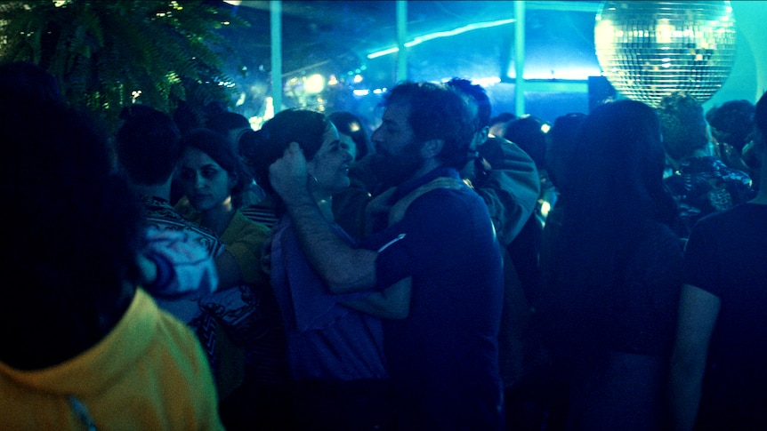 A woman and man hold each other closely on busy dancefloor in blue hue lit nightclub with disco ball.