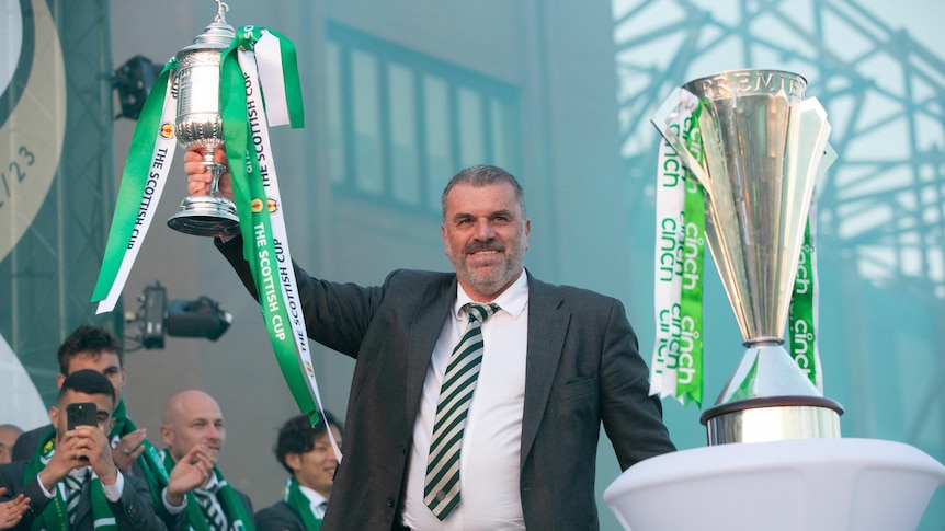 A smiling Ange Postecoglou stands on a stage holding a trophy with green and white ribbons, as another cup stands on a podium.,
