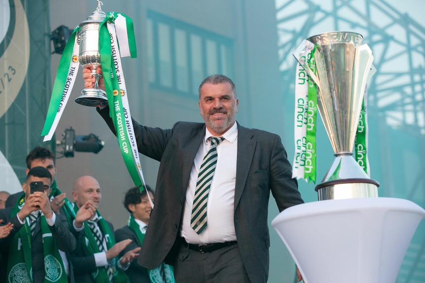 A smiling Ange Postecoglou stands on a stage holding a trophy with green and white ribbons, as another cup stands on a podium.,