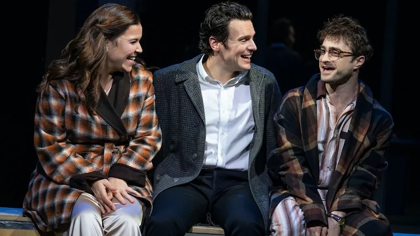 Three people sit on stage, smiling broadly at each other