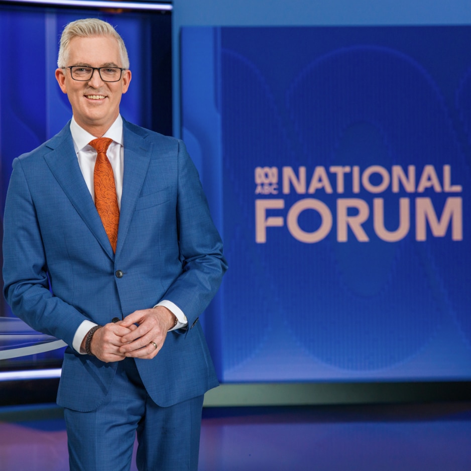 Smiling male TV host in a blue suit and orange tie stands beside a blue studio screen displaying ABC National Forum