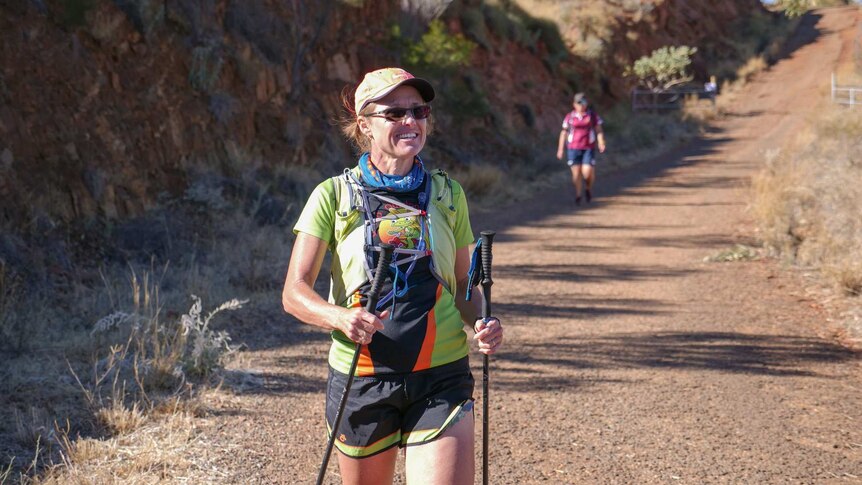 Woman in her late forties walks down a hill in mount isa with two hiking sticks in either hand.