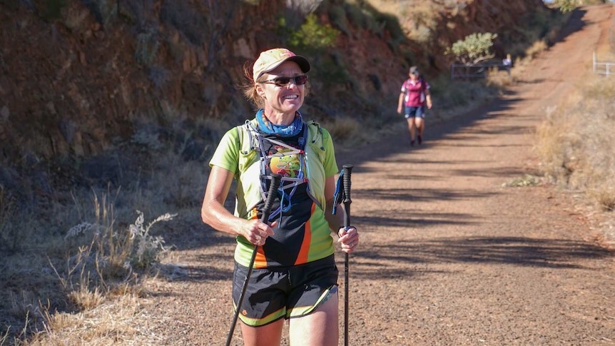 Woman in her late forties walks down a hill in mount isa with two hiking sticks in either hand.