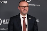 Andrew Barr wearing glasses and a suit speaks in front of a lectern.