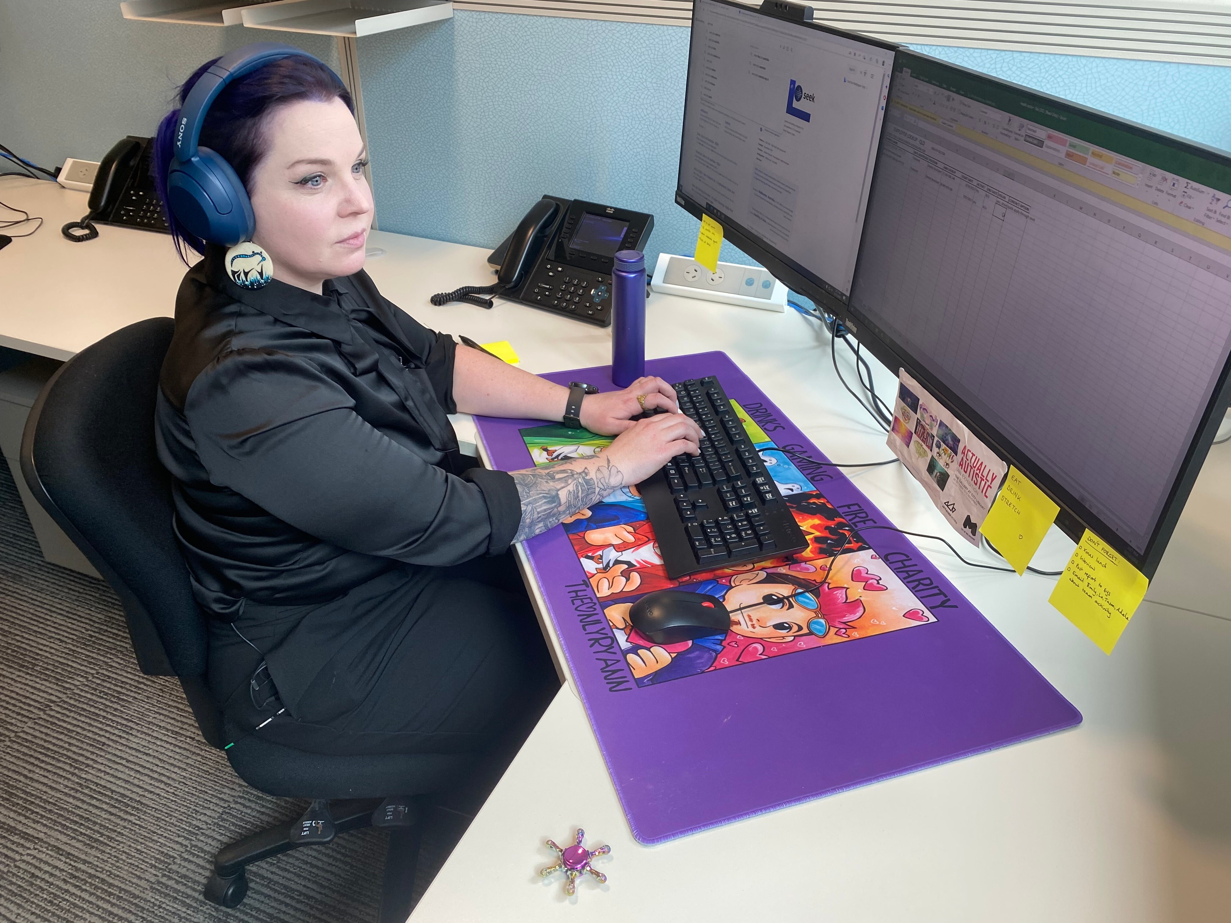 A woman sits at a computer, she has a black shirt and blue headphones on.
