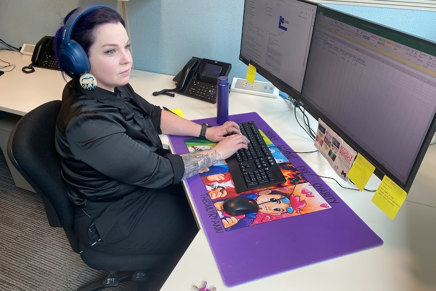 A woman sits at a computer, she has a black shirt and blue headphones on.