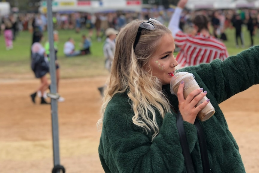 A side-on shot of a young blonde woman with glitter on her face drinking an iced coffee at a music festival.
