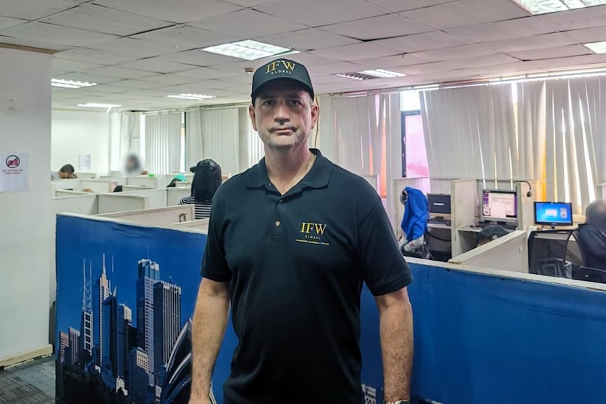 A man wearing a black shirt and black cap stands in an office.