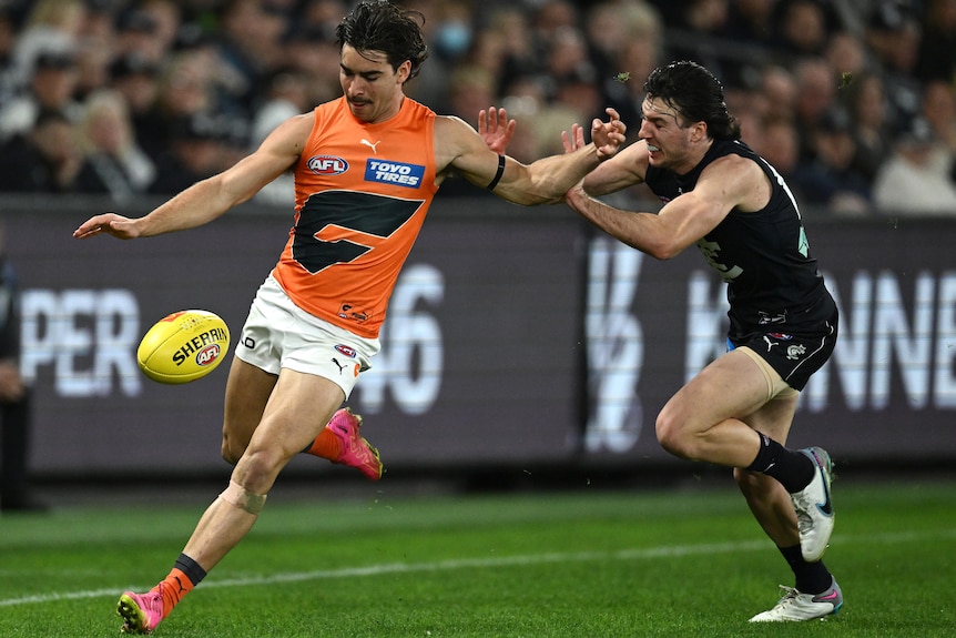 A GWS AFL player runs near the boundary and drops the ball to kick it goalwards as a Carlton defender closes in.