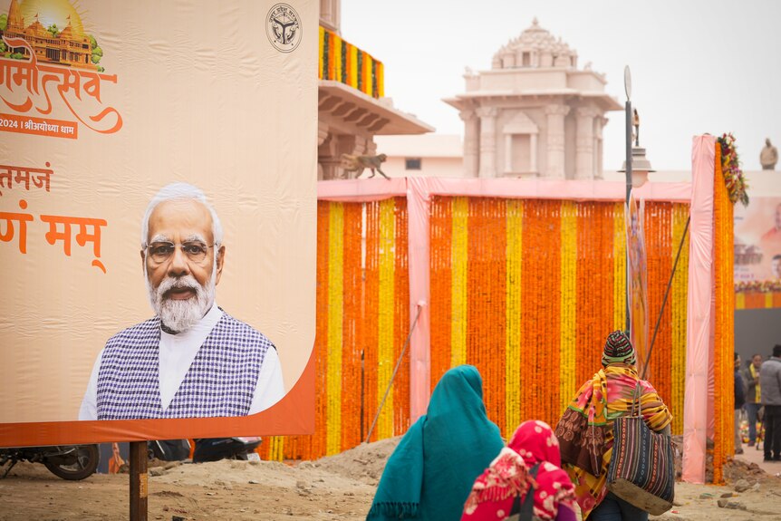 People walk past a billboard with Narendra Modi's face on it, a temple in the background