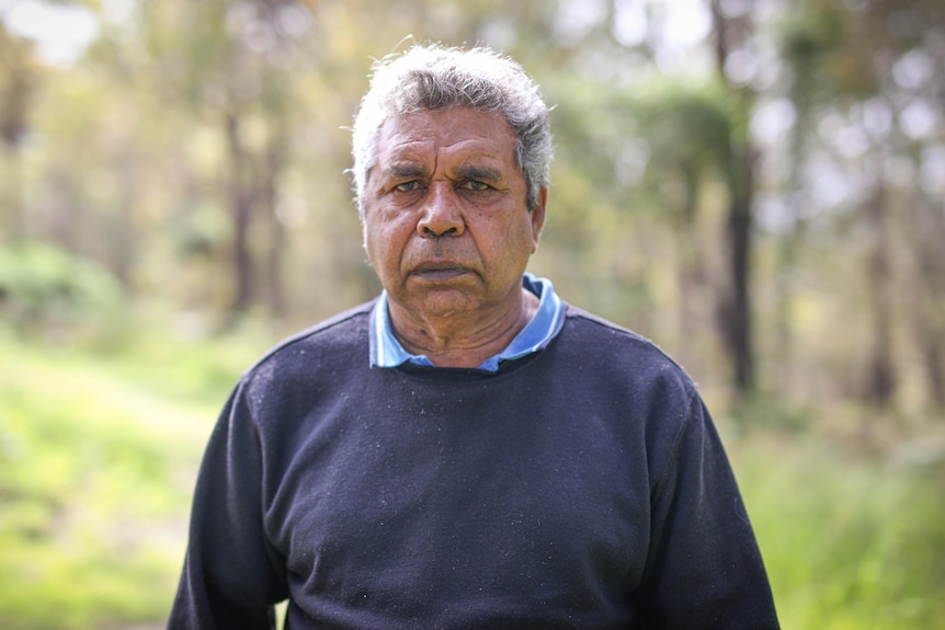A serious Aboriginal man in a blue shirt, jumper looks towards the camera, blurred greenery in the background. 