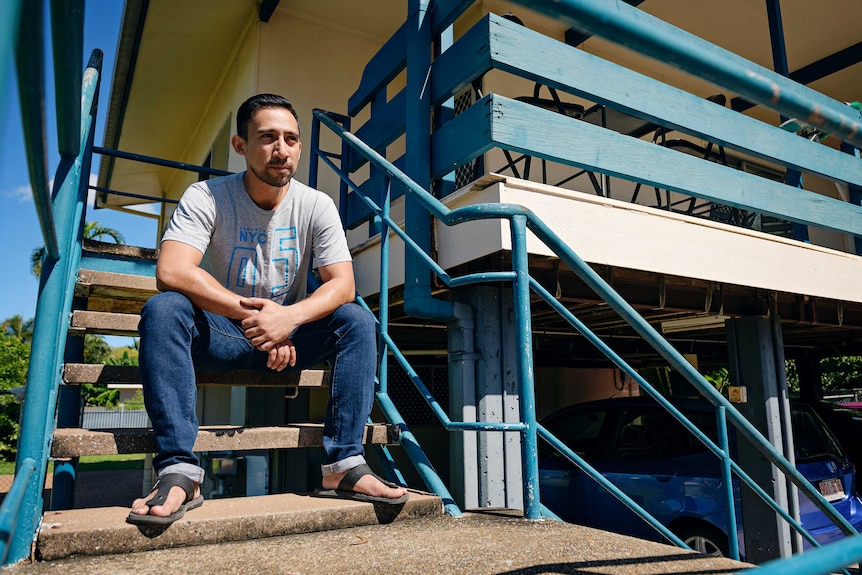 A man is seated on a stair case dressed in jeans and a t-shirt