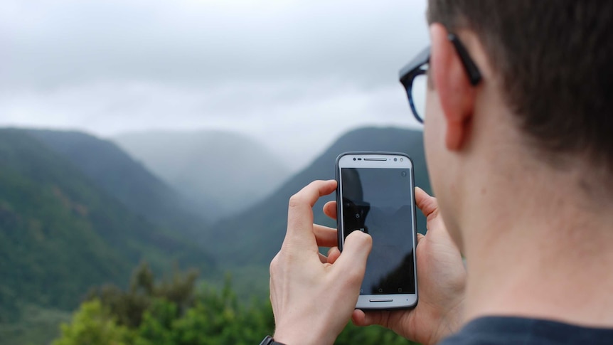 A man holds a phone in front of a natural landscape