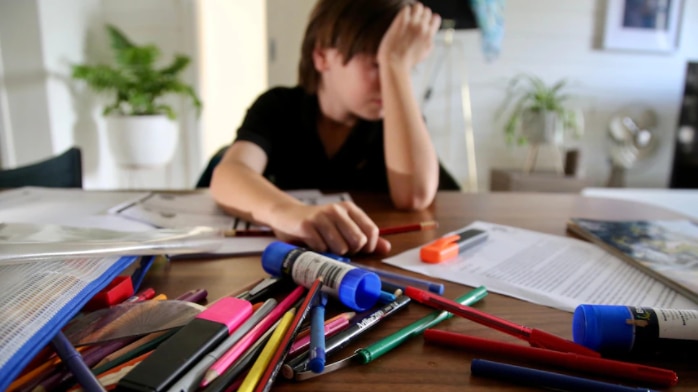 A child has his head in his hand as pens and pencils are strewn over workbook on table