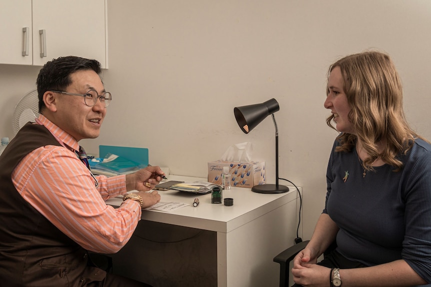 A man with short black hair, glasses, a pale pink shirt and grey waistcoast sits at desk talking to a woman with blonde hair.