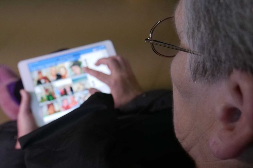 Old woman wearing glasses looking at facebook on an ipad on her lap, shot taken over her shoulder.