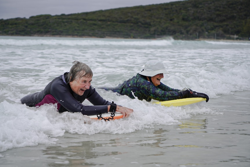 two elderly women wearing wetsuits lie on their boogie boards and catch a small wave in
