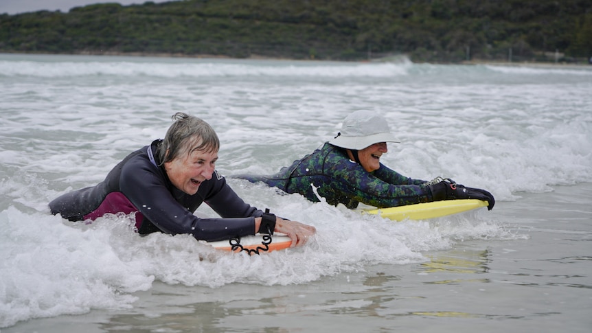 two elderly women wearing wetsuits lie on their boogie boards and catch a small wave in