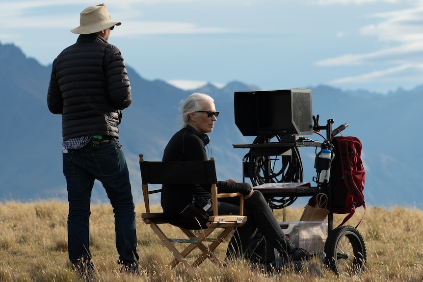 In a field in New Zealand, a middle-aged man stands behind a 60-something woman in a director's chair in front of a monitor