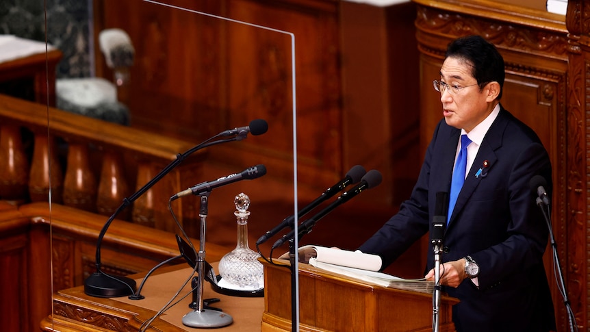 Fumio Kishida stands at a podium in a wood panelled room. 