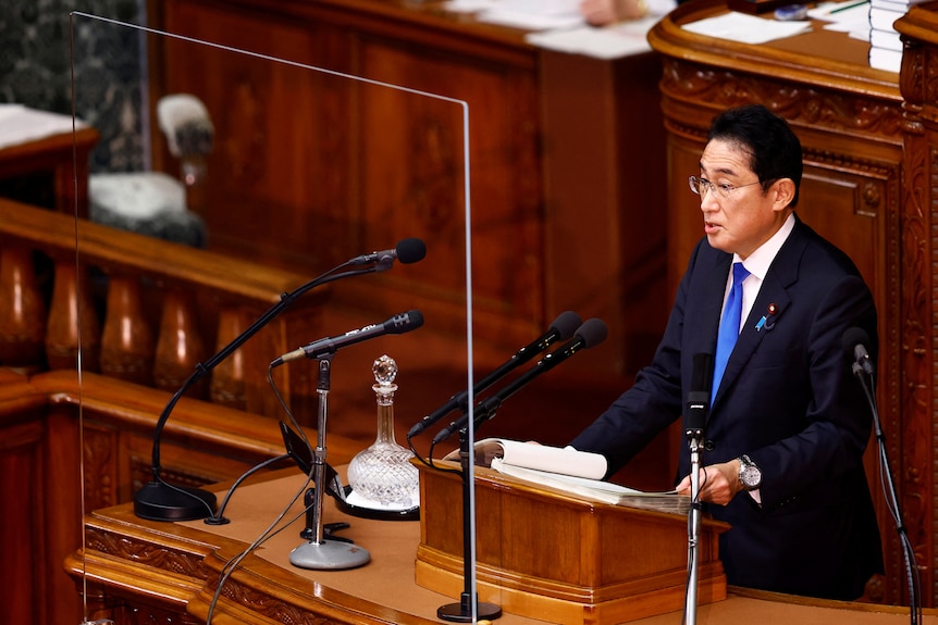 Fumio Kishida stands at a podium in a wood panelled room. 