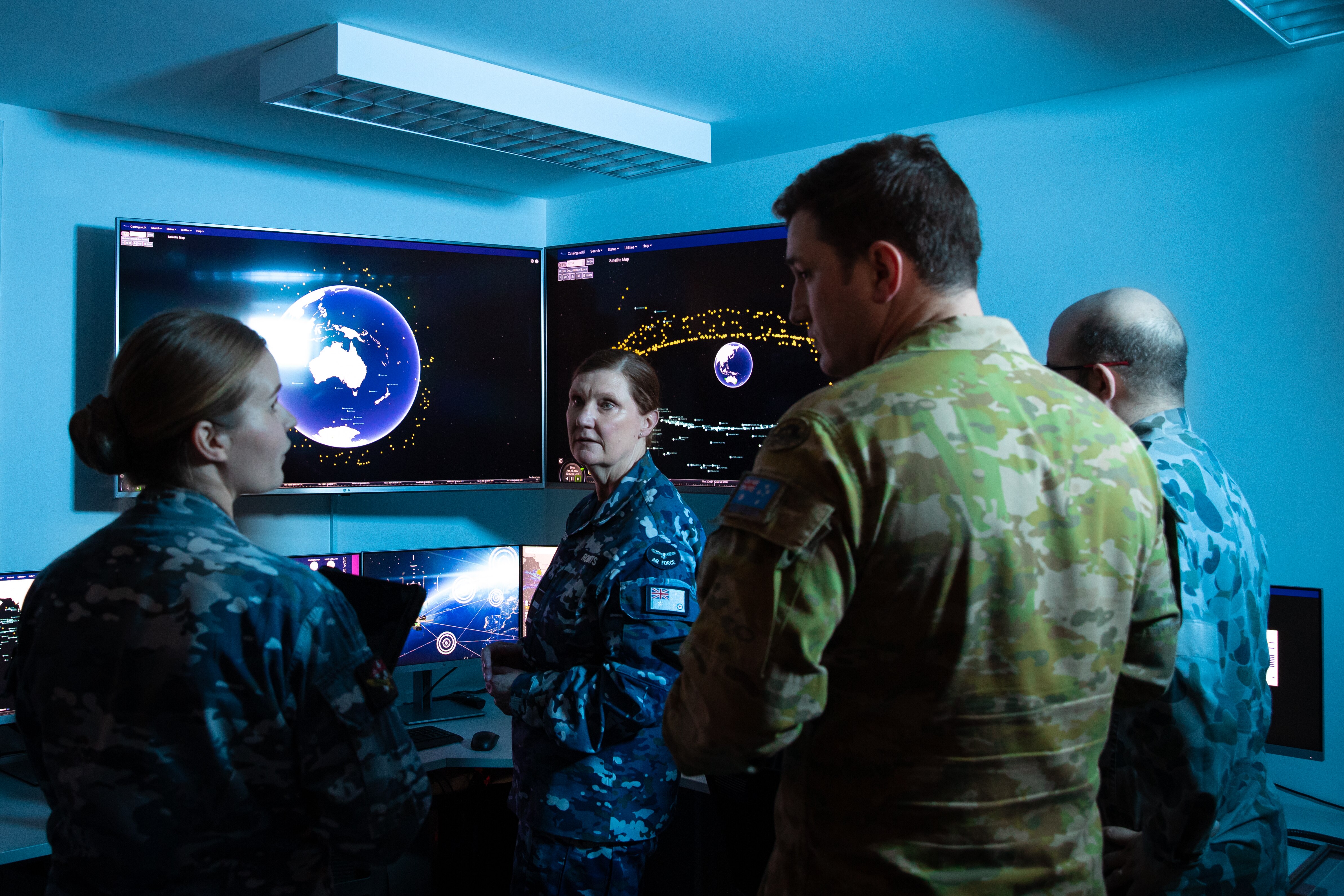 Four soldiers stand inside a dimly lit control room, with screens displaying planet Earth and orbiting satellites.