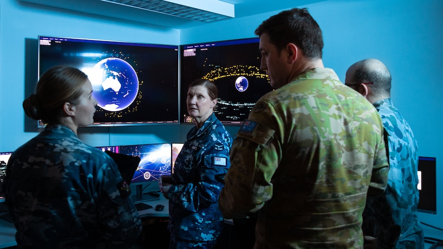 Four soldiers stand inside a dimly lit control room, with screens displaying planet Earth and orbiting satellites.