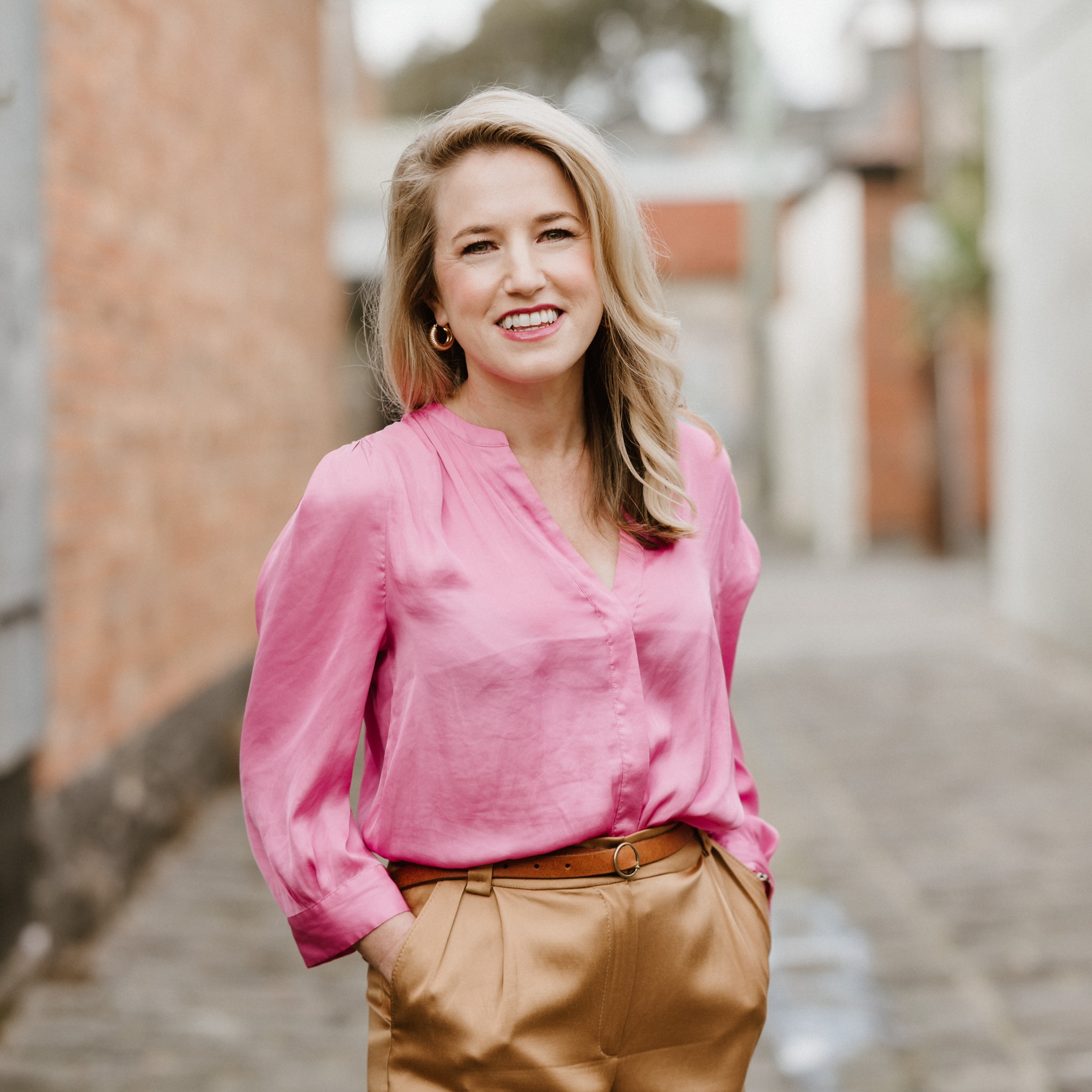 Uma foto profissional de Jacqueline Baulch tirada em uma rua de Melbourne. Ela veste uma blusa de linho rosa e calça camel.