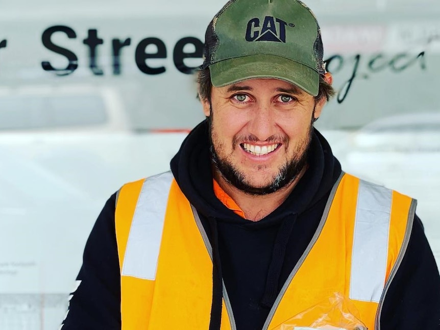 A young man in a green cap and orange high-vis vest smiling.