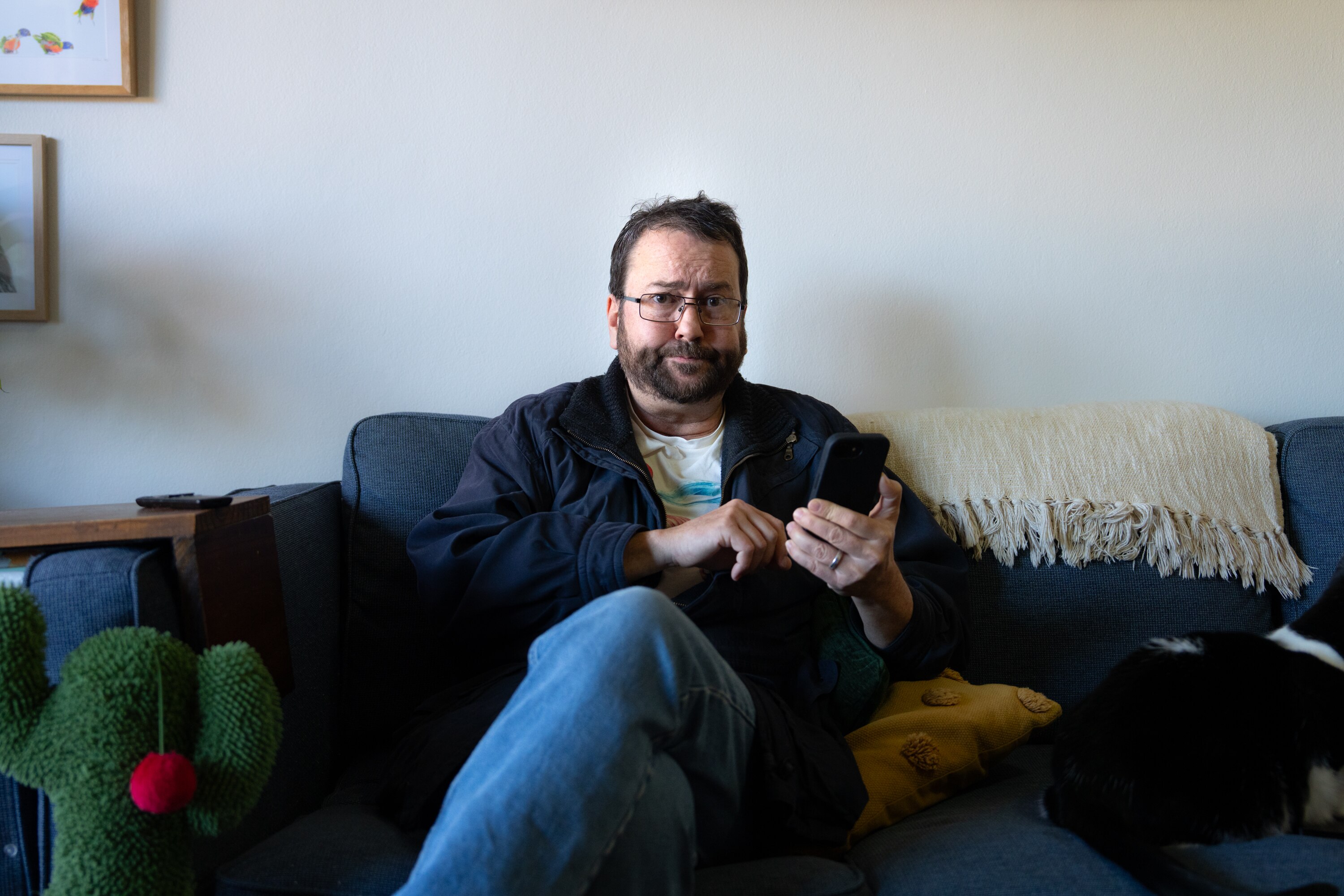 A man sitting on a couch, holding his phone and looking at the camera with a frown.
