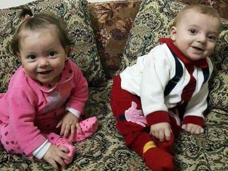 Young Syrian twin toddlers sit on a couch and look at the camera