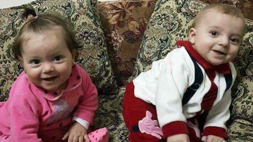 Young Syrian twin toddlers sit on a couch and look at the camera