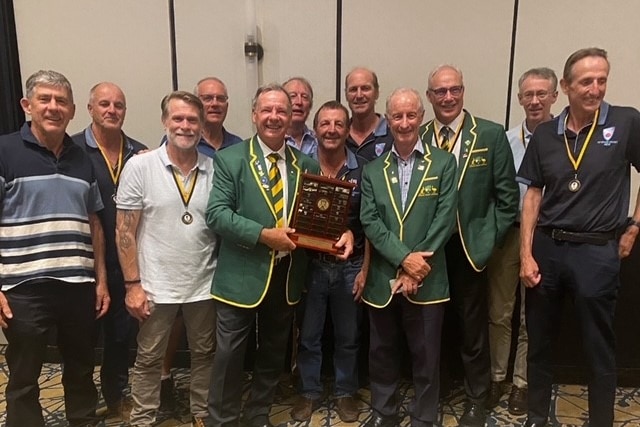 A group of men smile inside a room, holding a shield trophy.