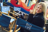 A woman sitting in a blue fold-out Big Little Lies chair, turned back towards the camera, holding up her left index finger.