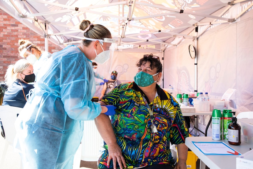 A worker in full PPE and a protective face shield speaks to a woman who is wearing a colourful NAIDOC shirt.