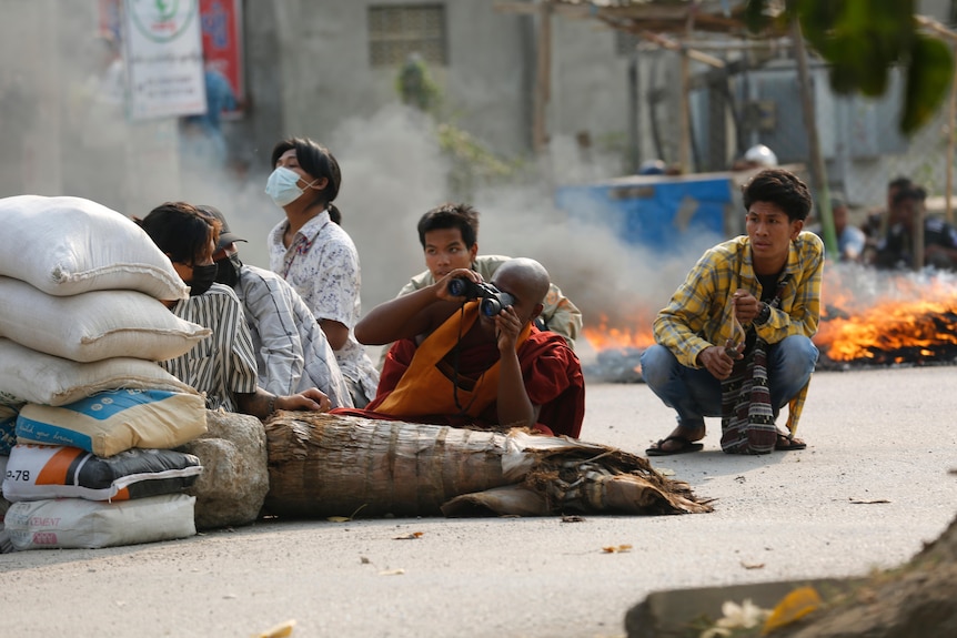 A Buddhist monk uses binoculars while others, including children, squat behind a road barricade.