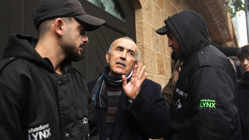two guards stand beside a man who is walking through a doorway while signalling with his hand a speaking to journalists