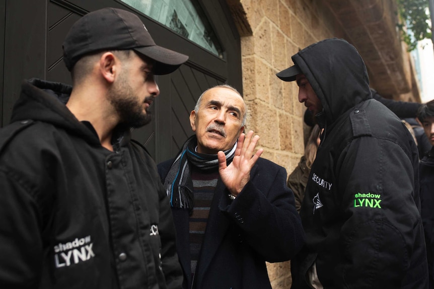 two guards stand beside a man who is walking through a doorway while signalling with his hand a speaking to journalists
