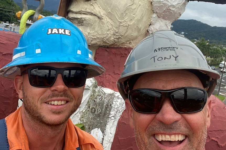 two men wearing hard hats and sunglasses pose for a selfie with a giant statue