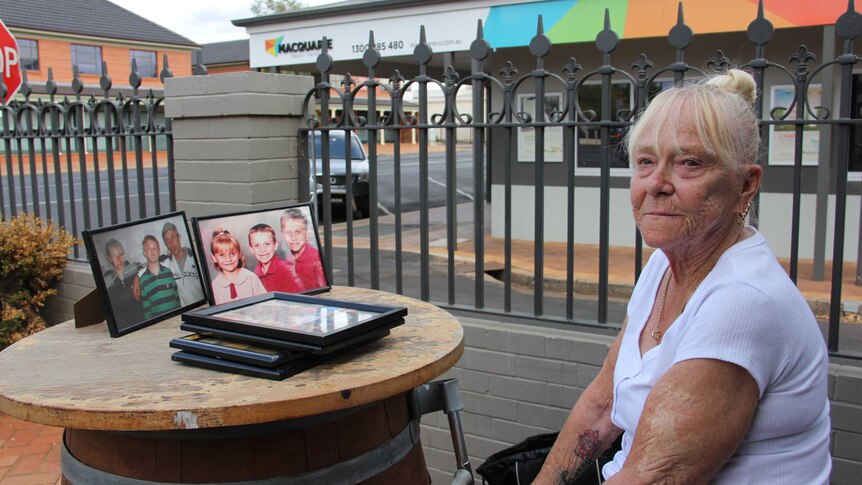 A woman sits at a table with framed photographs of a young children and teens.