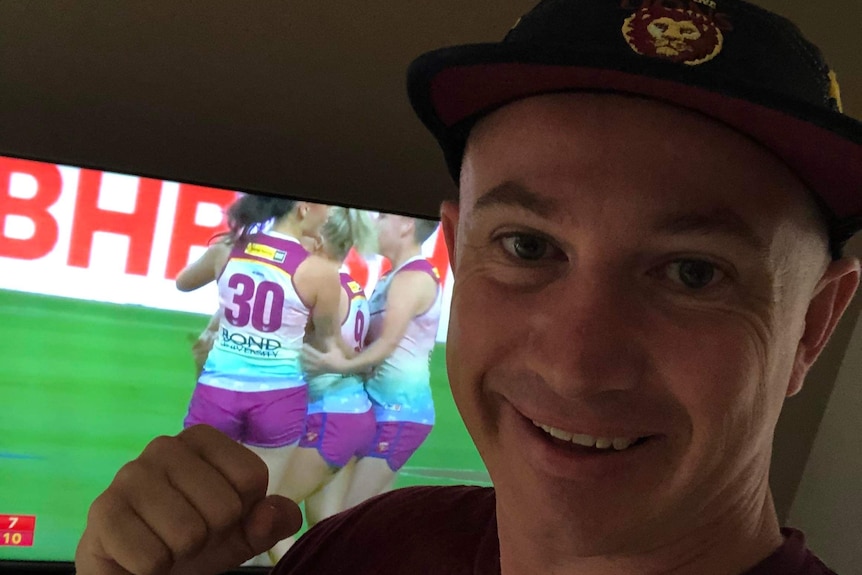 Andrew Dower stands in front of a tv playing an AFLW game and clenches his fist.