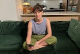 A young woman sits on a couch and looks into camera with a serious expression.