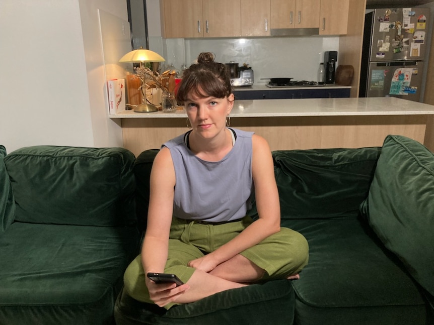 A young woman sits on a couch and looks into camera with a serious expression.