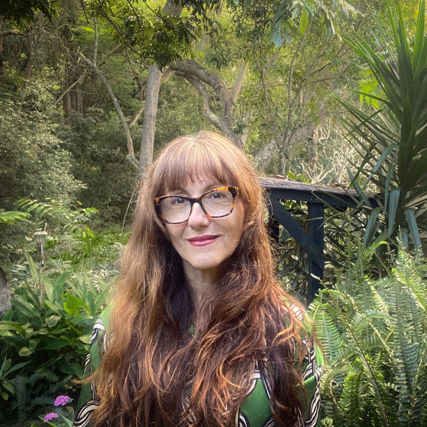 A woman with long red hair and glasses stands in amongst a tropical garden of greenery.