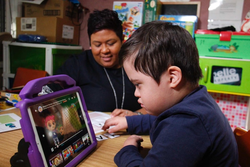 Victor Taschke plays on a device in the foreground, while his mother Divina sits beside him.