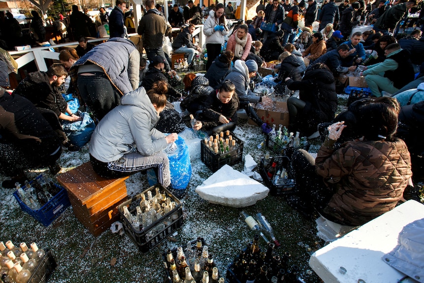 A crowd of people in winter clothes huddle around empty glass bottles, preparing Molotov cocktails.