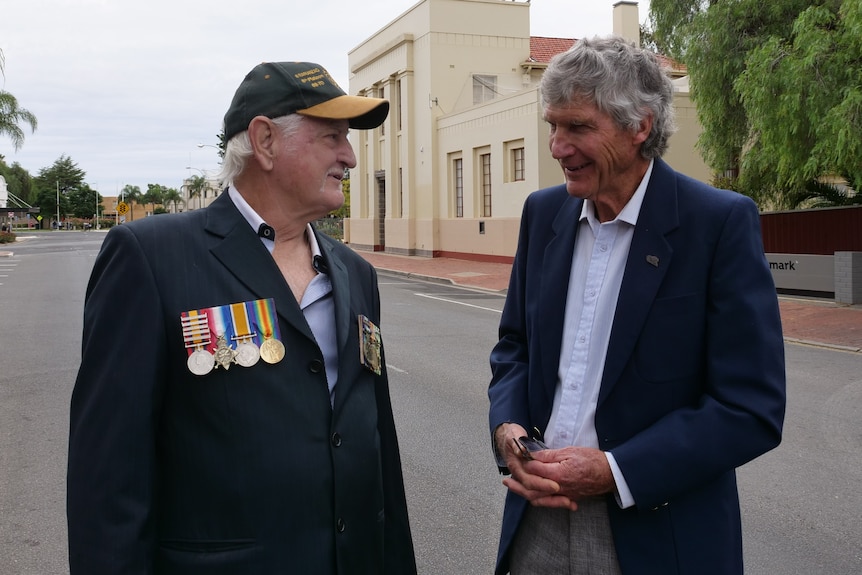 Two men talk to each other, they are on the road, one is wearing a hat and medals.