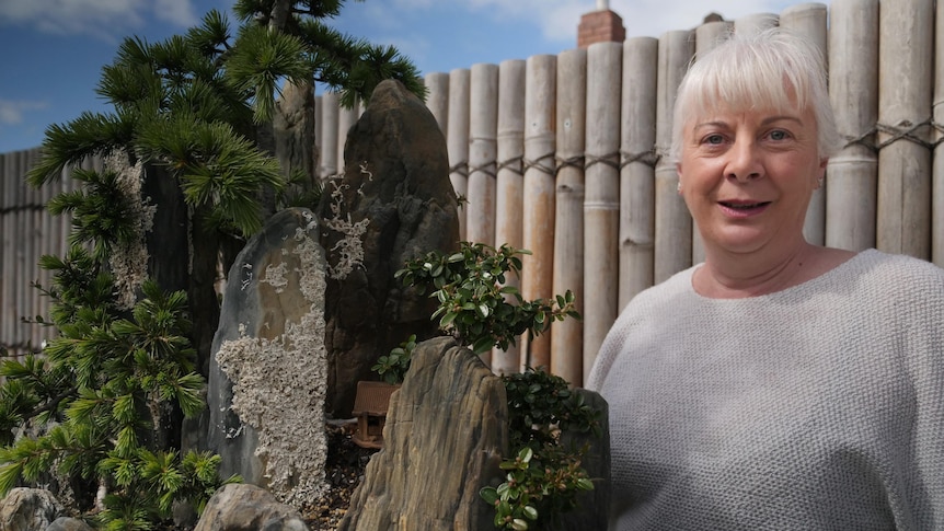 A woman standing next to a bonsai display, including a number of different small trees and rocks.