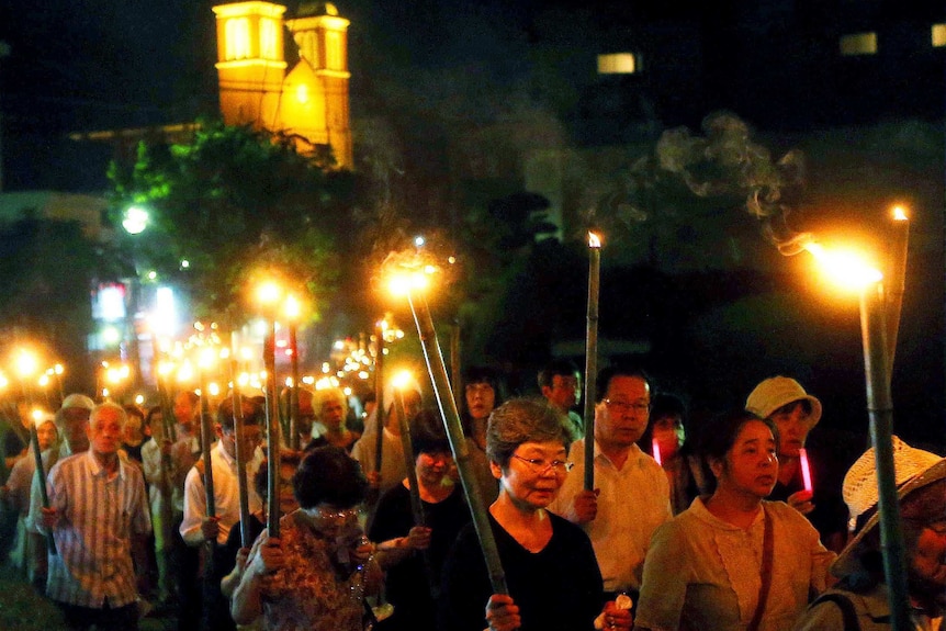 A line of people hold bamboo torches in a line at night, in front of a Cathedral in the background.