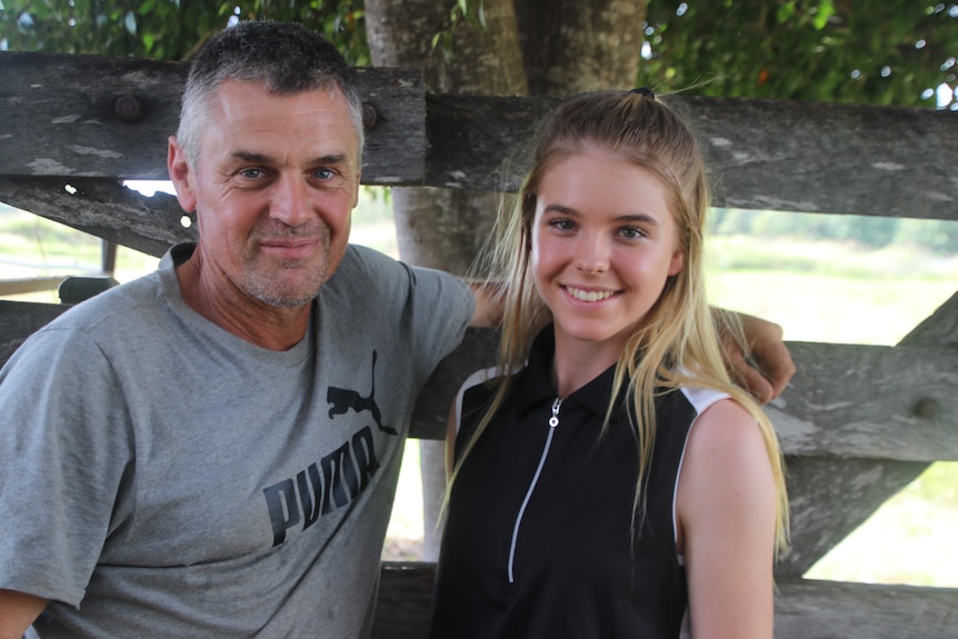 Father on the left in a grey shirt standing beside his daughter in a black shirt in front of an old gate